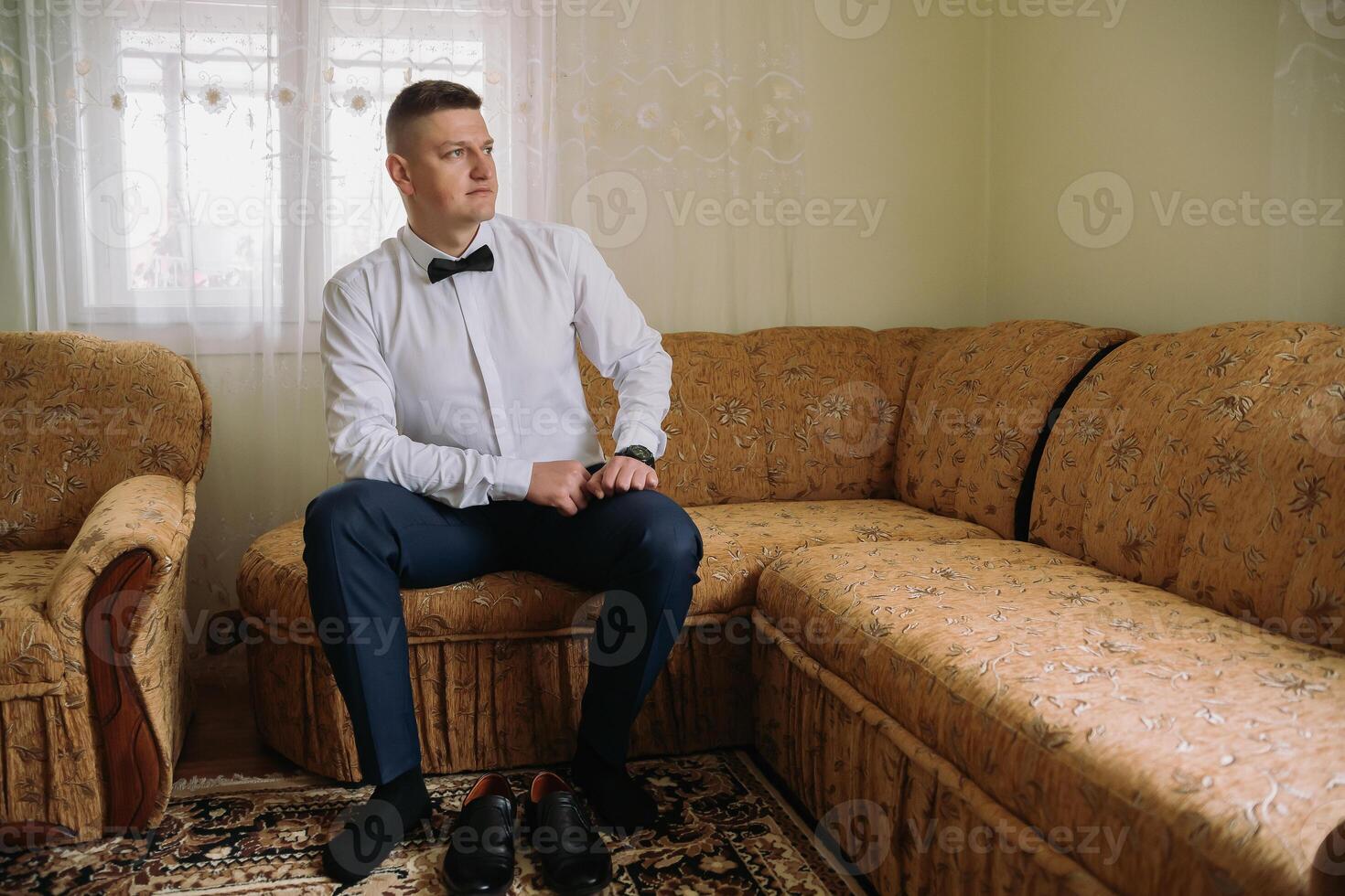 A man in a suit and bow tie sits on a couch photo