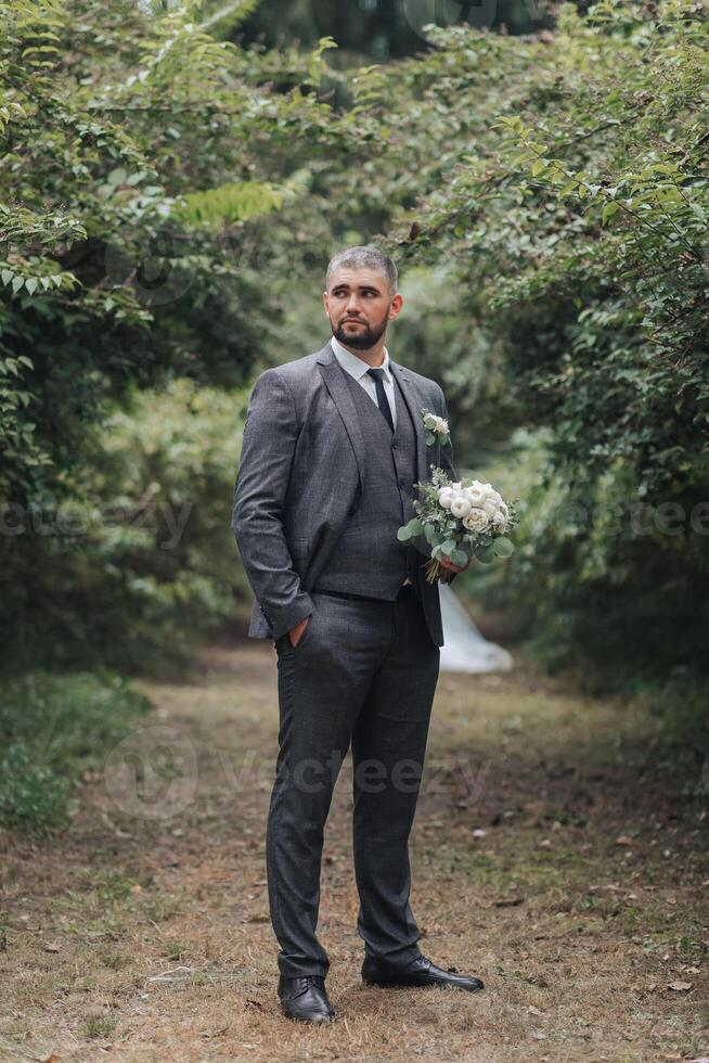 A man in a suit and tie stands in a forest with a bouquet of flowers photo