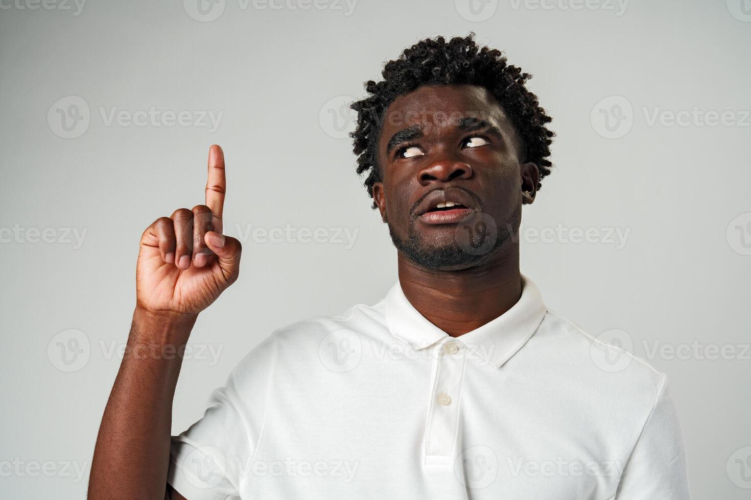 Young African Man in Casual Attire Pointing Upwards With a Smile Against a Grey Background photo