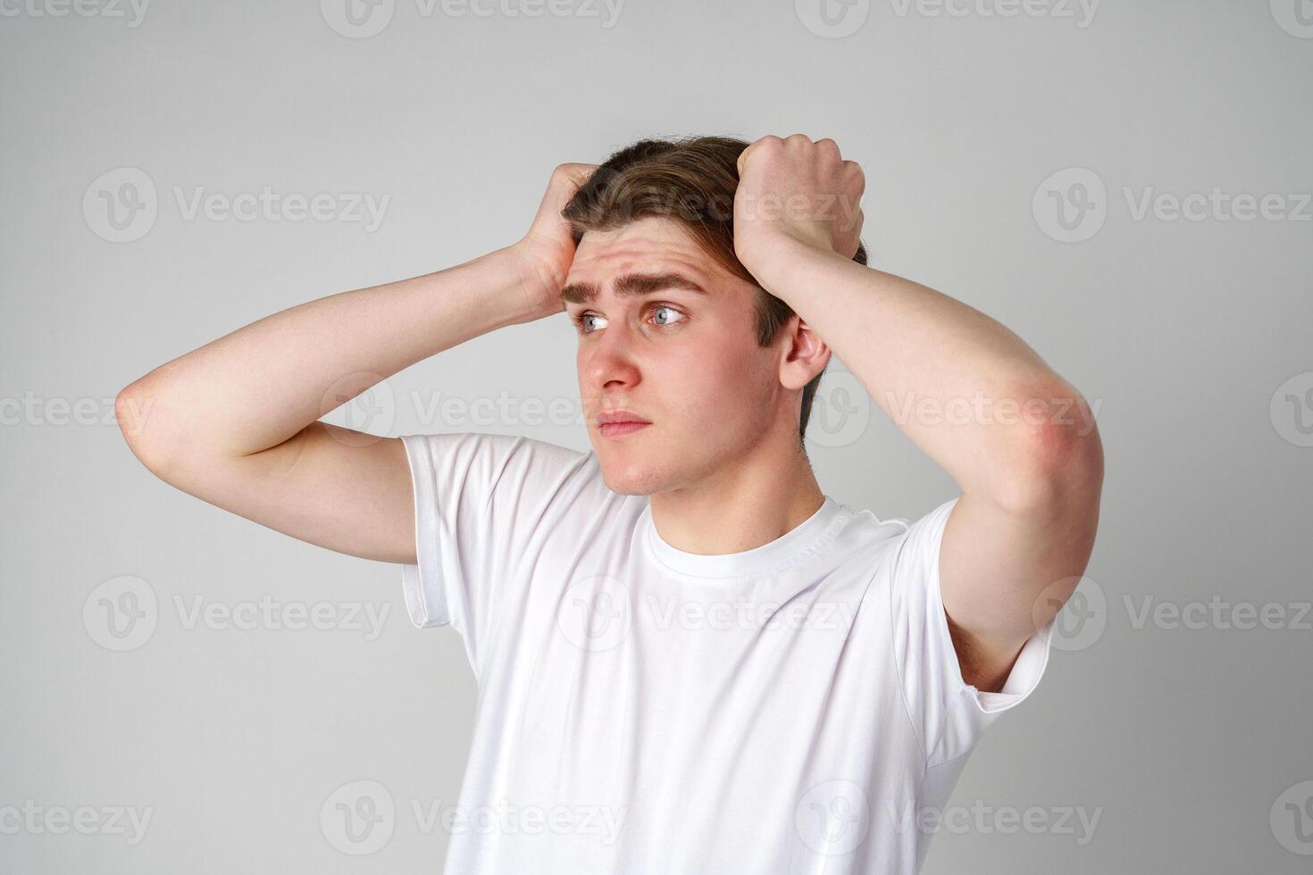 Young Man In White T-Shirt Looking Worried With Hands On Head Against Plain Background photo