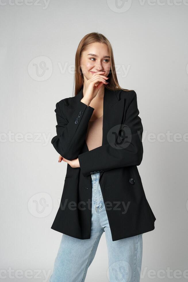 Young Woman in Casual Blazer and Jeans Smiling Against a Grey Background photo