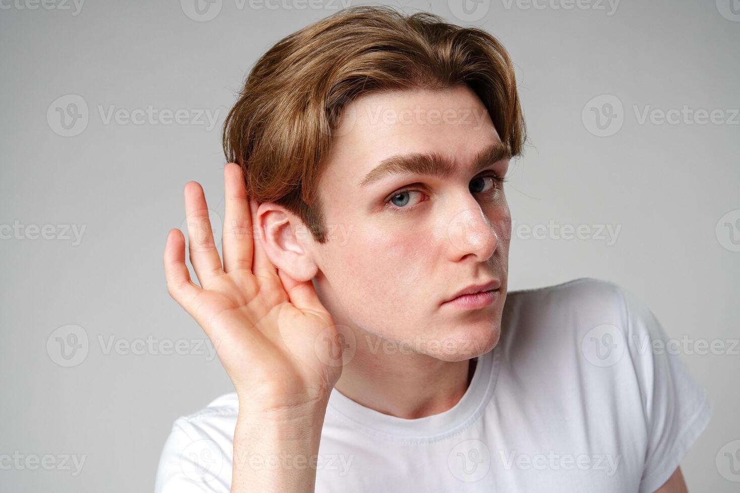 Young Man Placing Hand Behind Ear to Listen More Attentively Against a Neutral Background photo