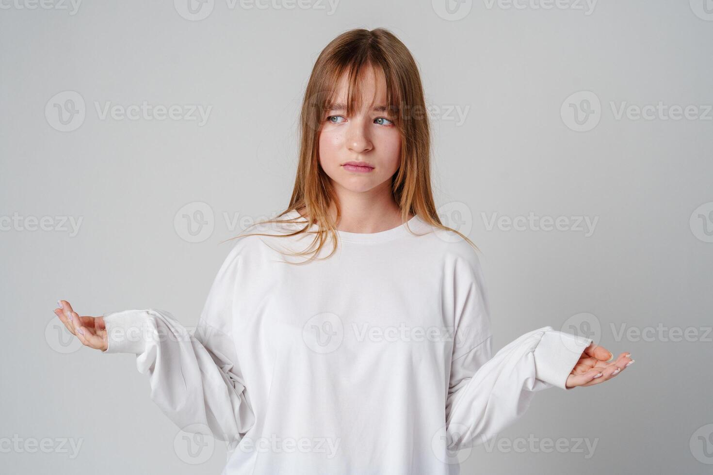 A young girl wearing a white long-sleeve shirt expressing confusion in a minimalistic gray studio setting photo