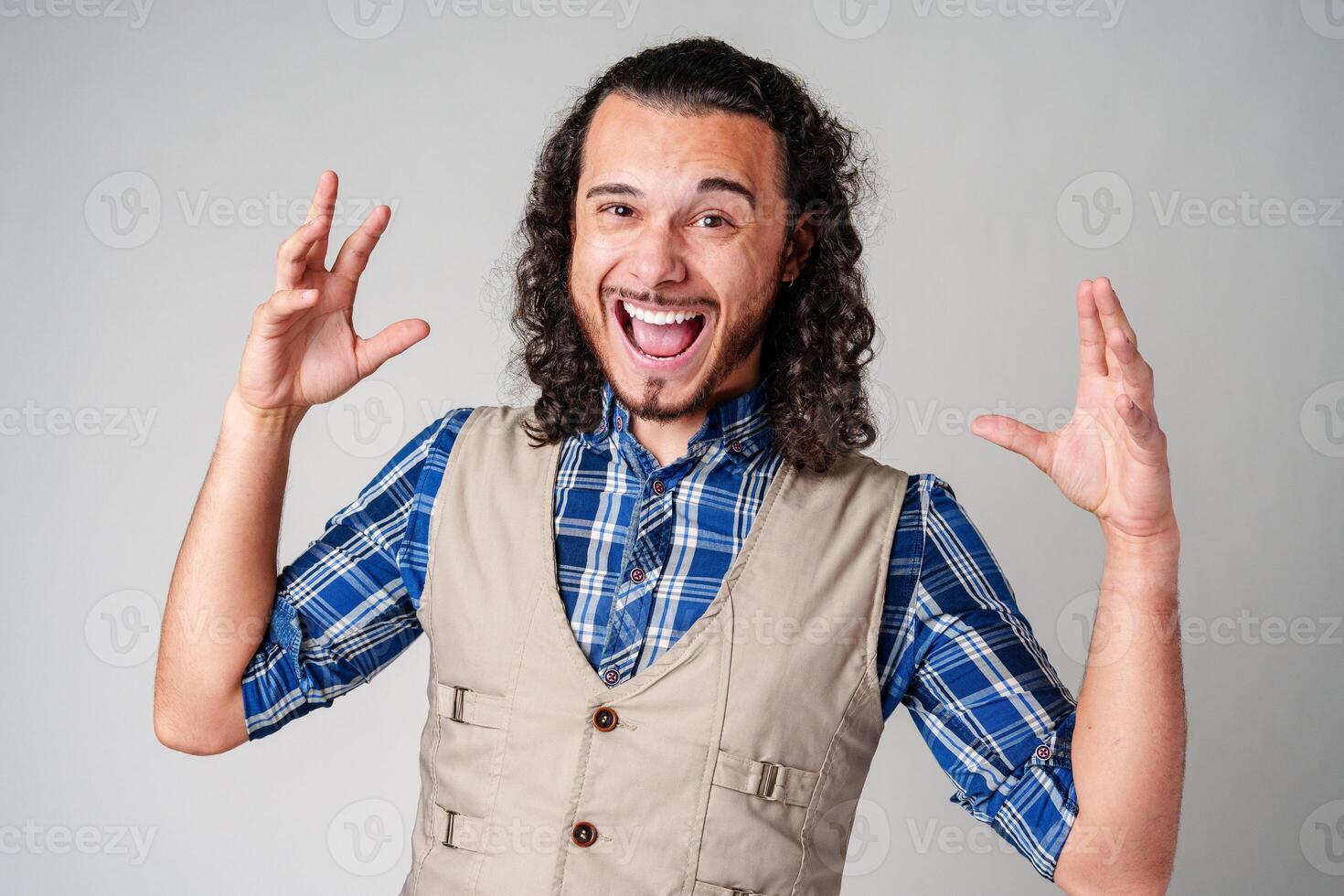 A man with curly hair excitedly gesturing while wearing a plaid shirt and vest against a plain background photo