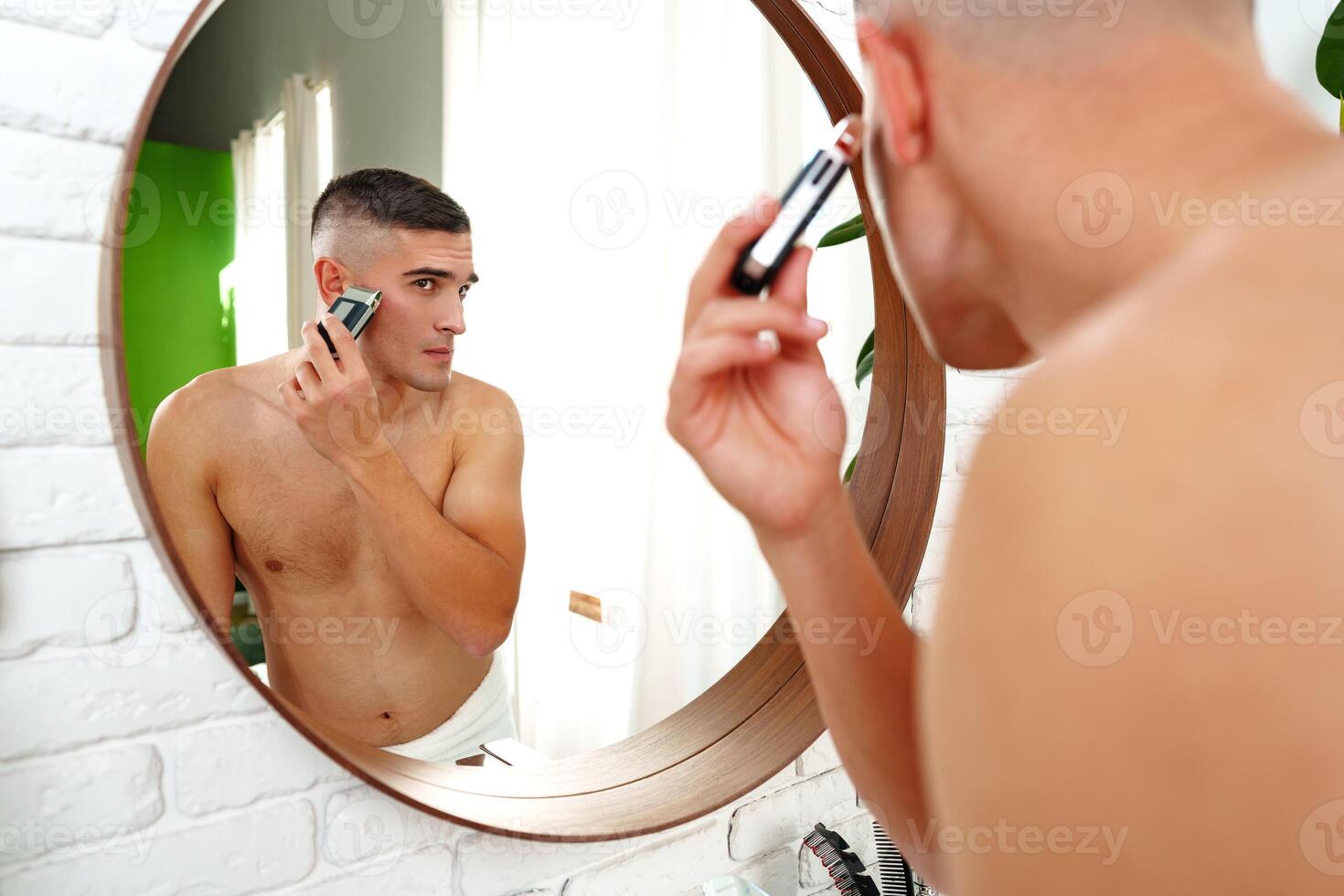 Handsome young man shaving face with electric razor on white background photo