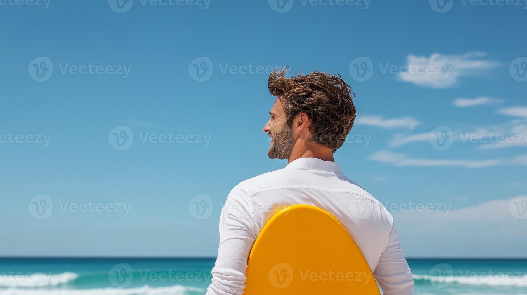 A man with a surfboard enjoys a sunny day at the beach, looking out at the waves and clear blue sky in summer photo