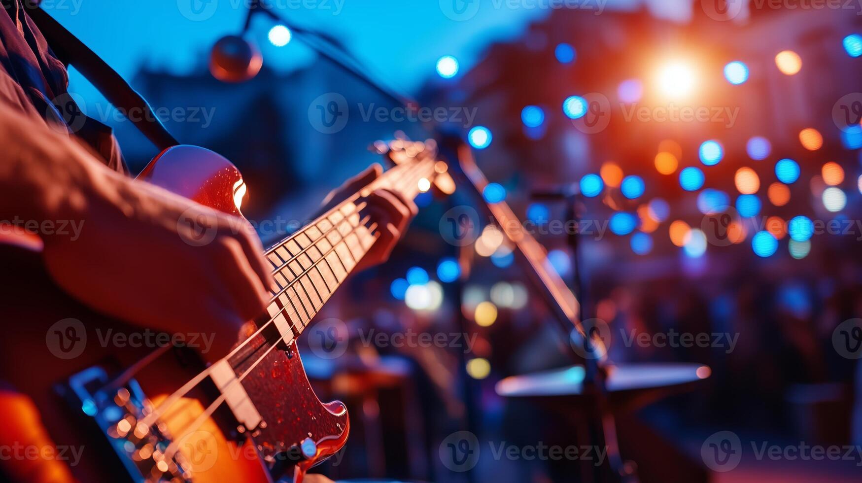 A musician plays a bass guitar on stage during a vibrant outdoor concert at dusk with colorful lights illuminating the crowd photo