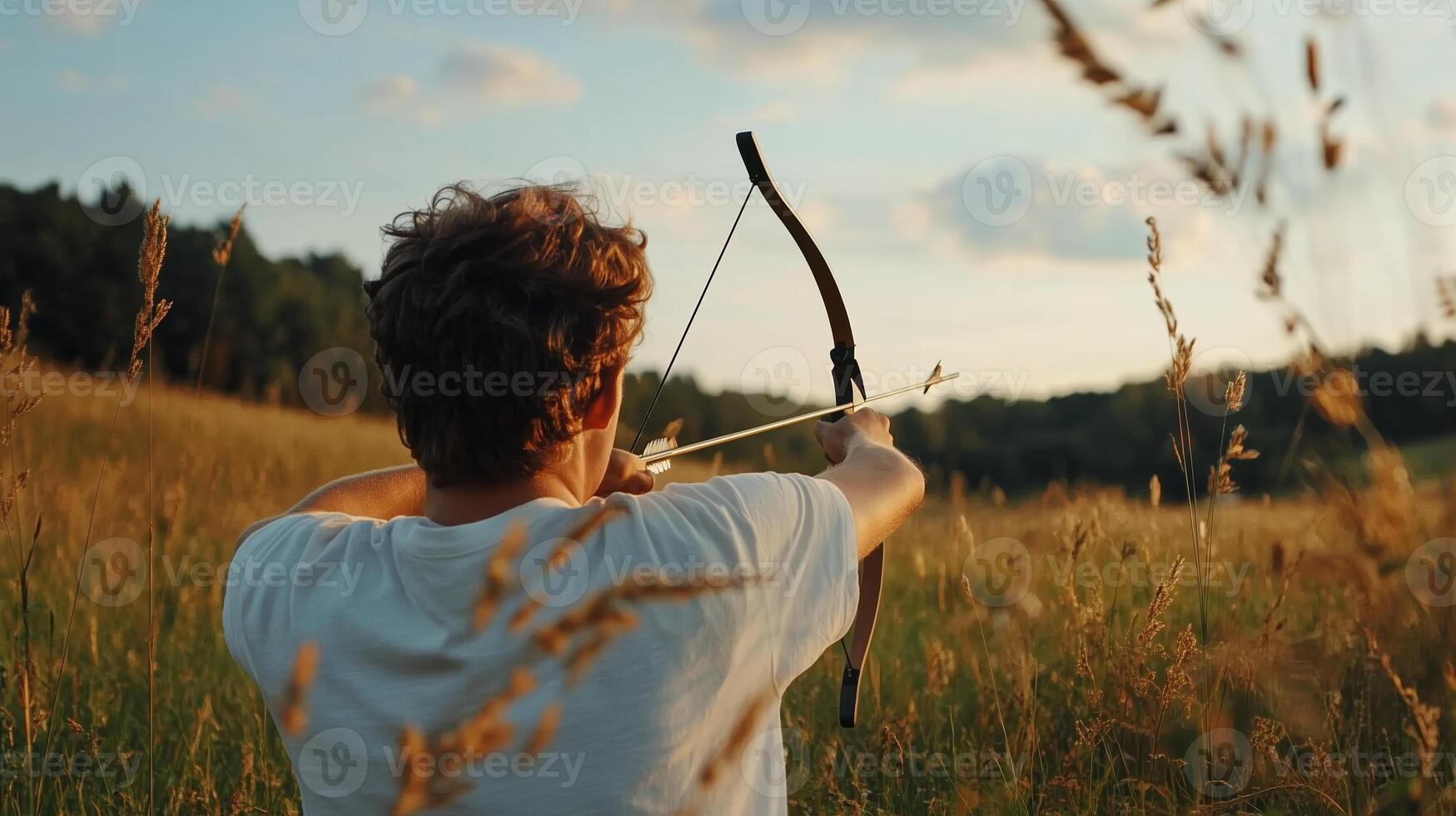 A young archer practicing with a bow at sunset in a grassy field surrounded by trees photo