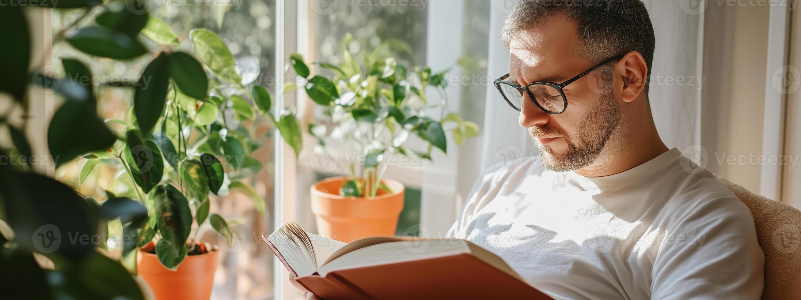 A man reading a book while sitting by the window among potted plants in a bright, cozy living room photo