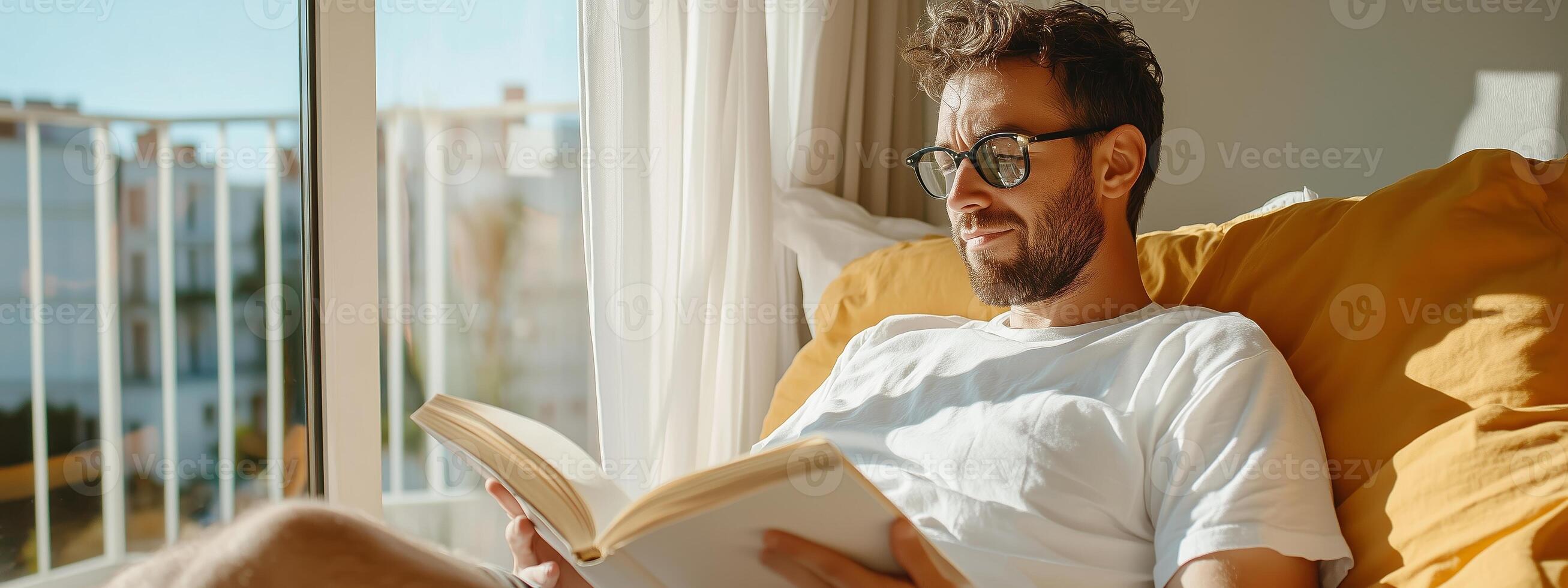 A man relaxes in a sunlit room while reading a book on a cozy yellow sofa in a modern apartment photo