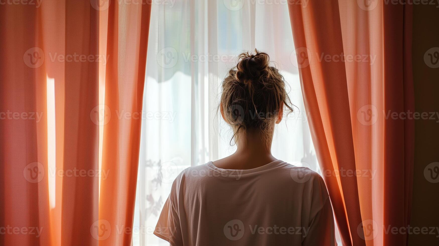 A person gazes out a bright window through sheer curtains in a cozy indoor space during the daytime photo