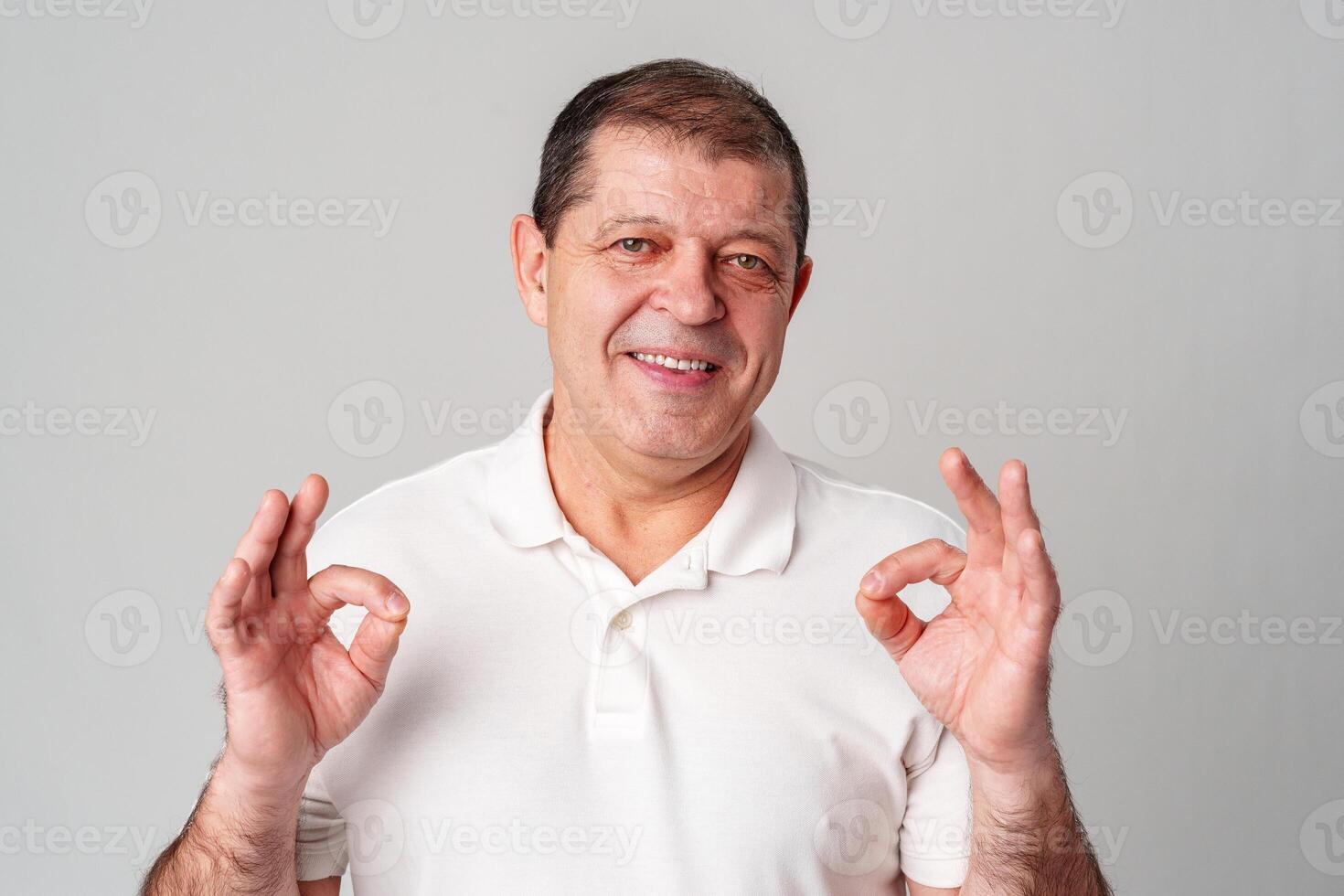 A man in a white shirt poses with a cheerful expression showing OK hand gestures in a well-lit indoor setting photo