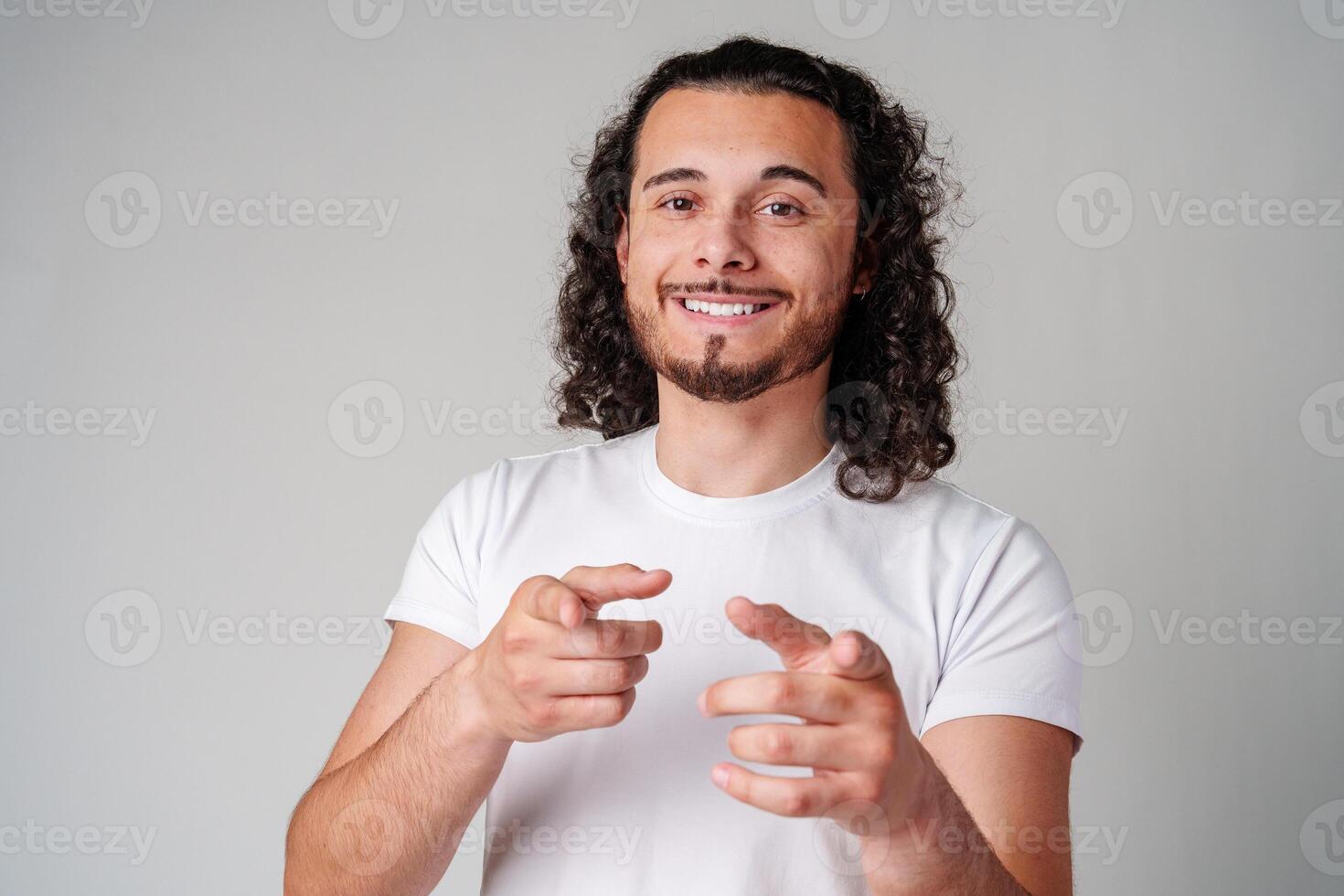 A smiling young man with curly hair playfully pointing directly at the viewer in a light-colored setting photo