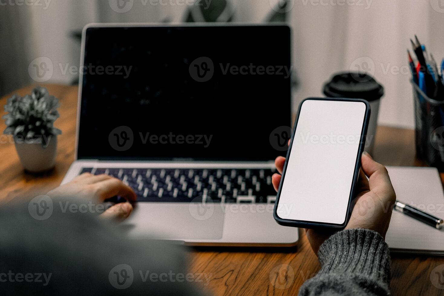 Person using a smartphone while sitting at a desk with a laptop, suggesting multitasking or mobile communication. The screen of the smartphone is blank. photo