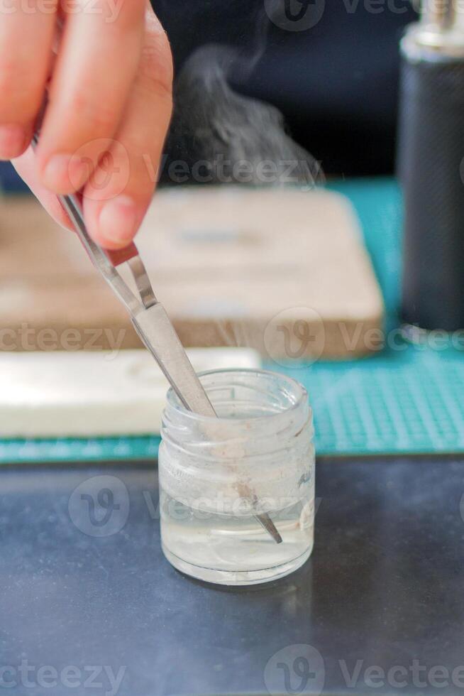 Close-Up of hand of jeweler holding tweezers with silver ring in glass jar with water and smoke. Workplace of jeweler. photo