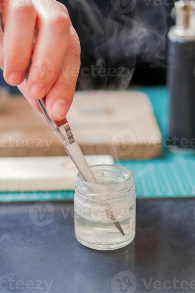 Close-Up of hand of jeweler holding tweezers with silver ring in glass jar with water and smoke. Workplace of jeweler. photo