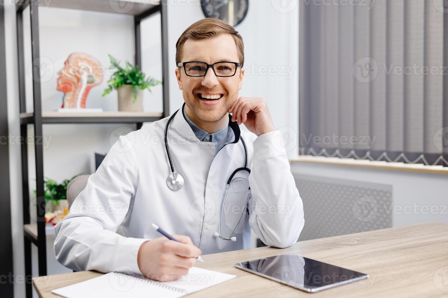 Portrait of young doctor wearing glasses and white uniform with stethoscope, speaking and consulting patient online, looking at camera, making call, sitting at table in office photo
