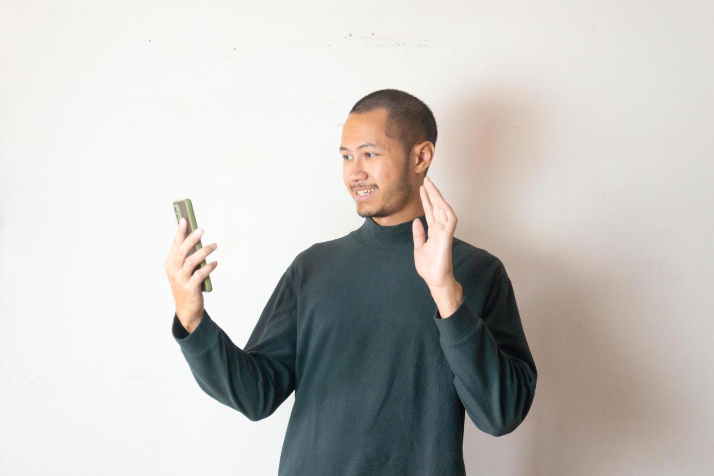 Young men wearing green sweater waving hand to smartphone calling friend on isolated white background photo