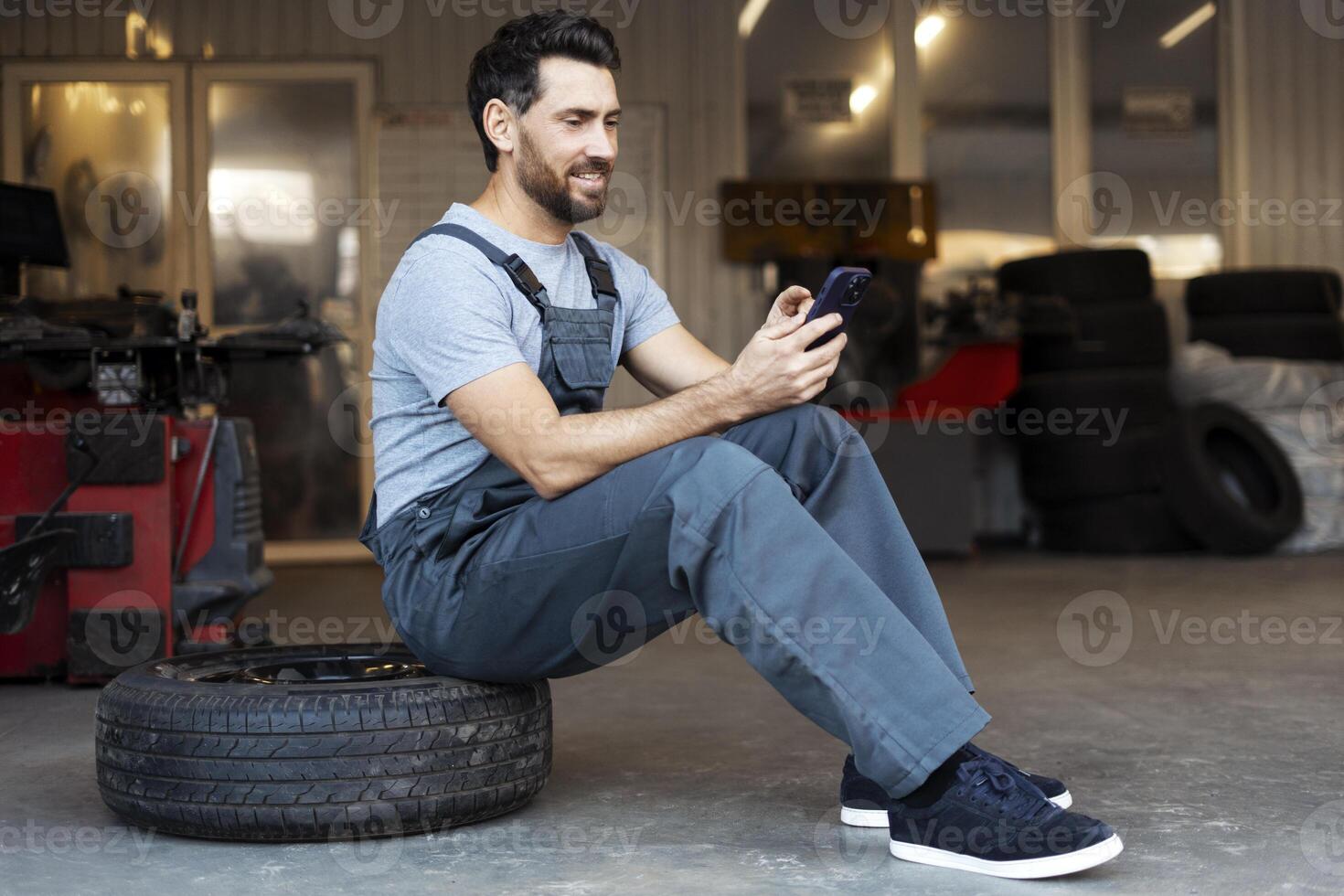 Mechanic sitting on tire browsing on mobile phone in auto repair shop photo