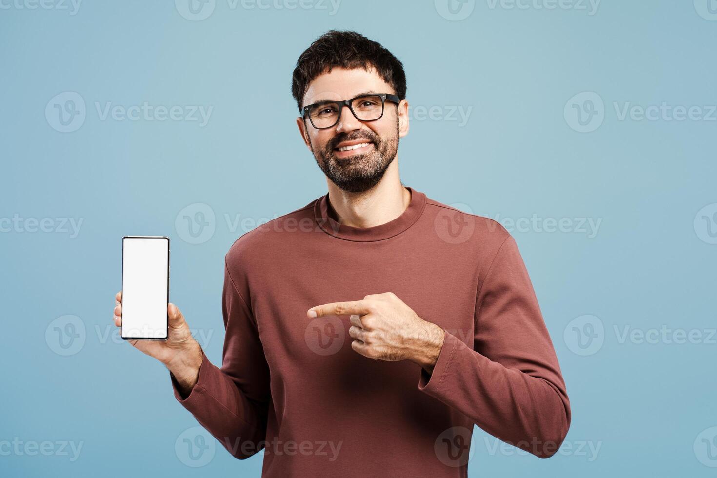 Smiling Hispanic man holding mobile phone with empty screen, pointing finger on mockup, copy space photo