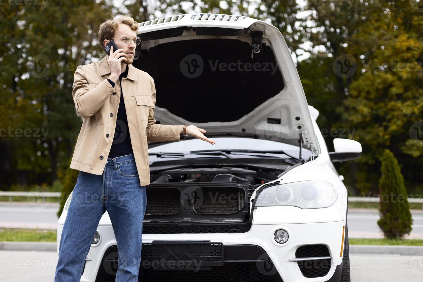 Confused driver calling mechanic on mobile phone near broken car with open hood photo