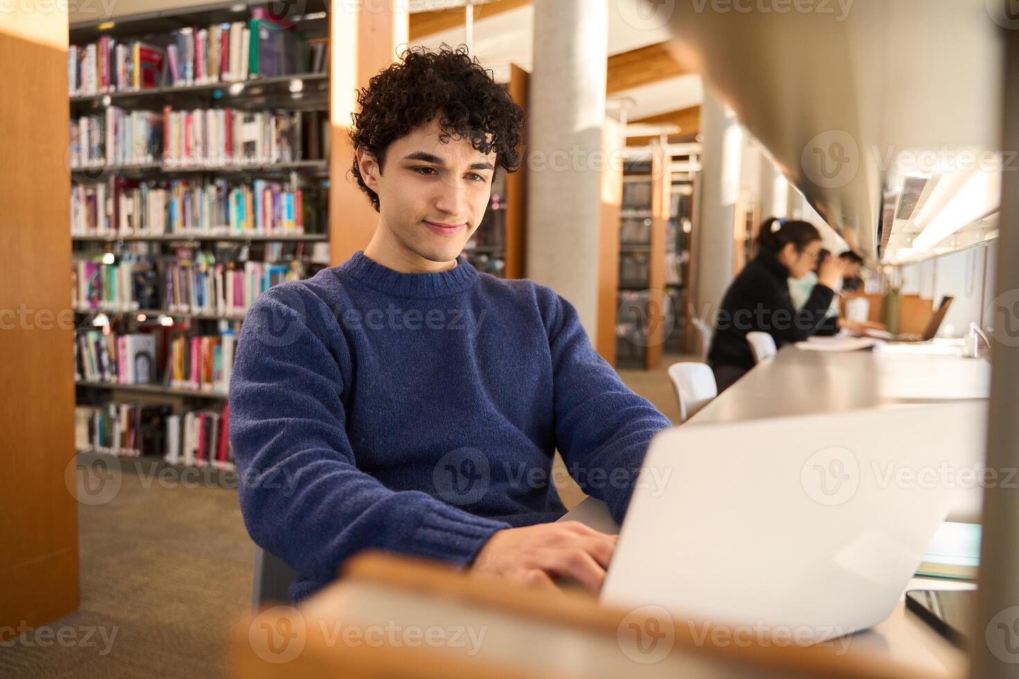 Young man working on laptop sitting at desk in busy public library photo