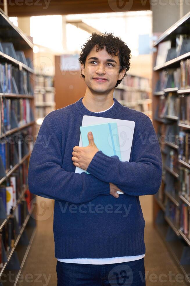 Thoughtful student holding books and laptop in library photo