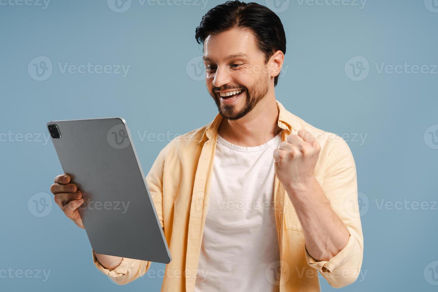 Excited bearded man celebrating success while holding digital tablet isolated on blue background photo