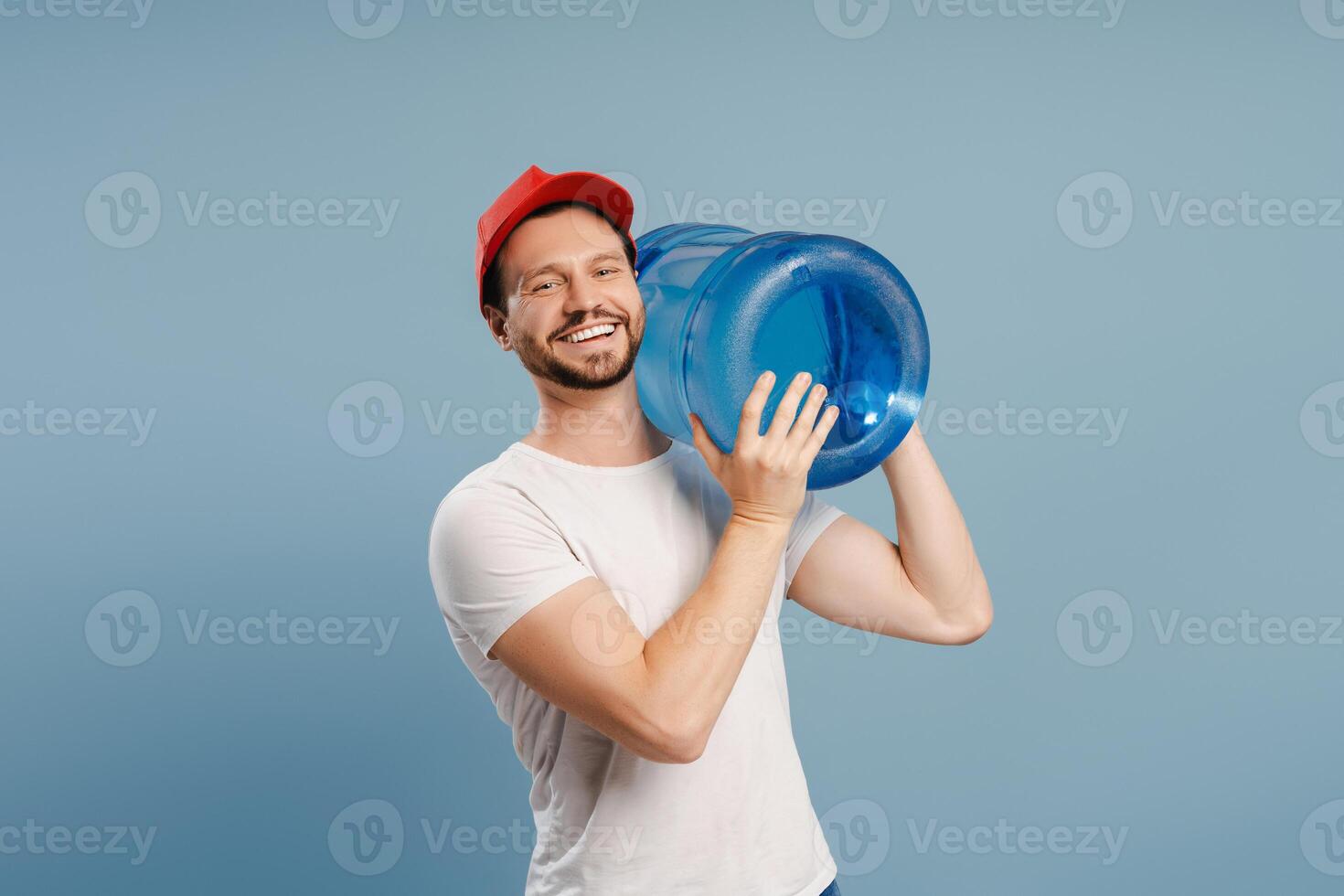 Delivery man holding heavy water bottle smiling at work posing on blue background. Service concept photo