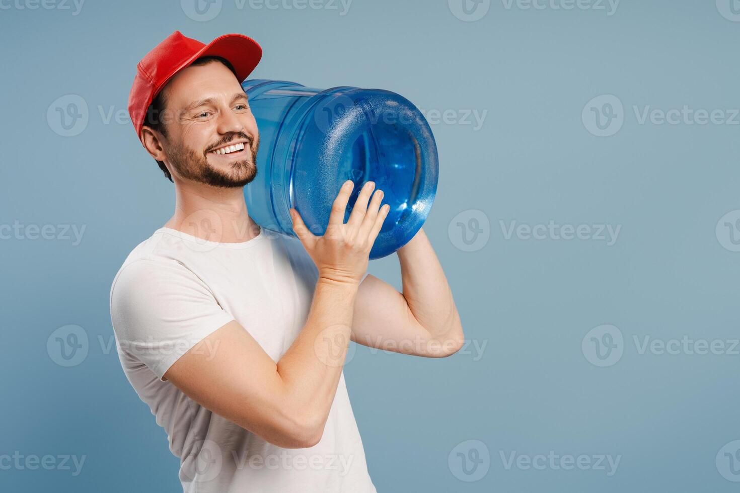 Delivery man smiling and carrying heavy water bottle on blue background photo