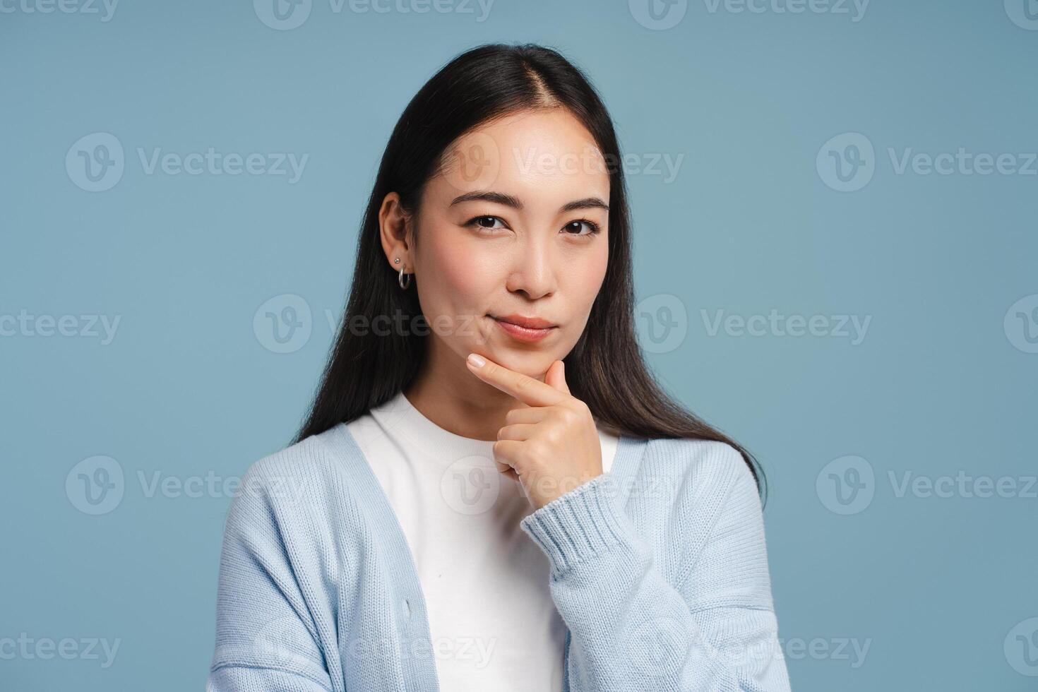 Pensive, young Asian woman touching chin and thinking looking at camera on blue background photo