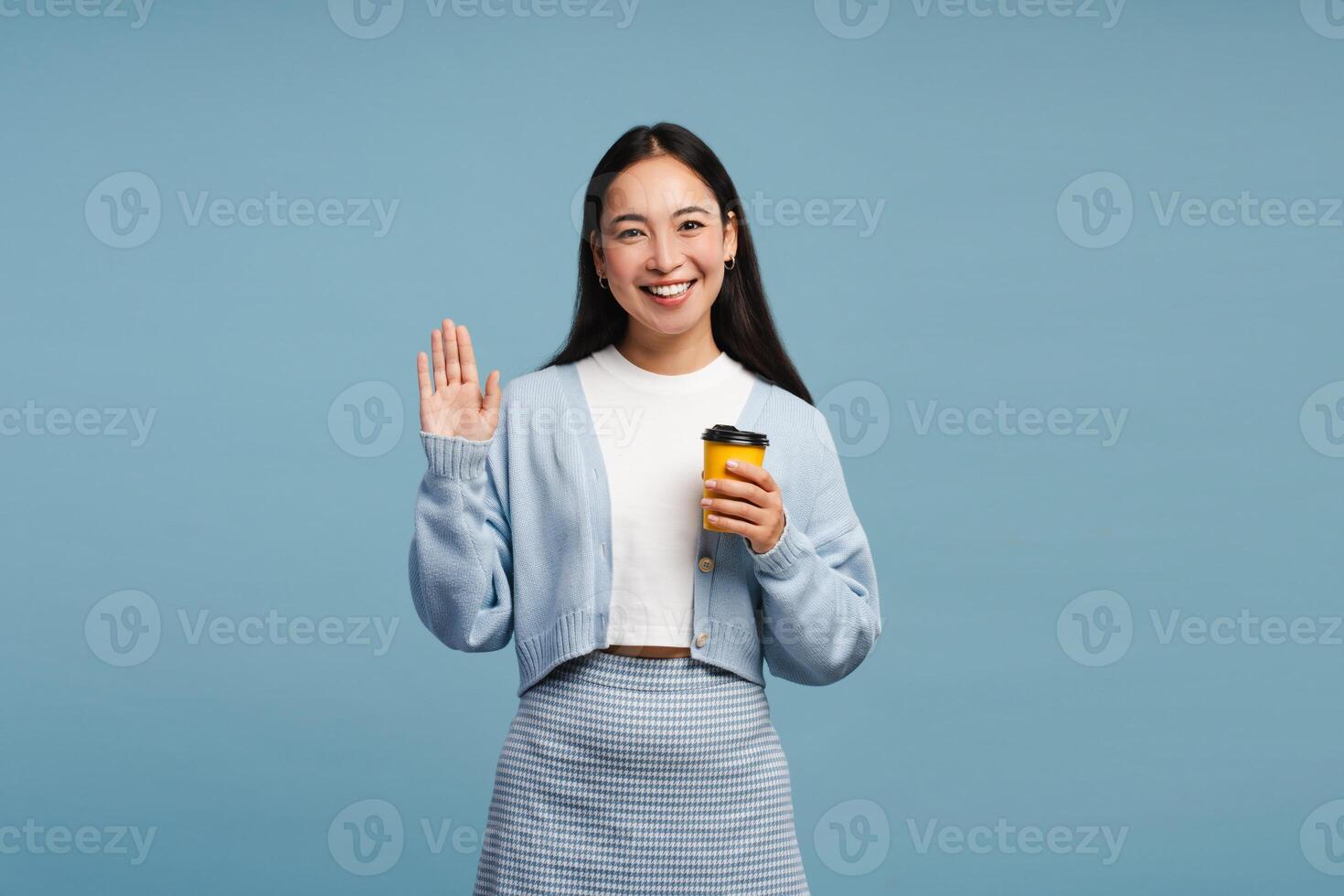 Smiling young Asian woman holding coffee cup and waving hand on blue background photo