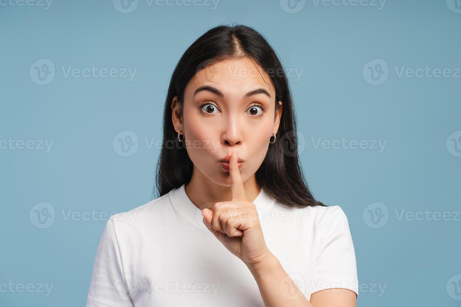 Surprised Asian woman gesturing, showing sign for silence with finger on lips on blue background photo