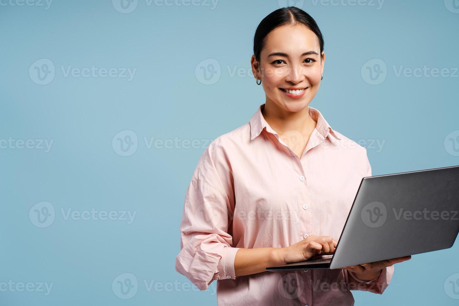Smiling attractive Asian woman typing on laptop looking at camera isolated on blue background photo