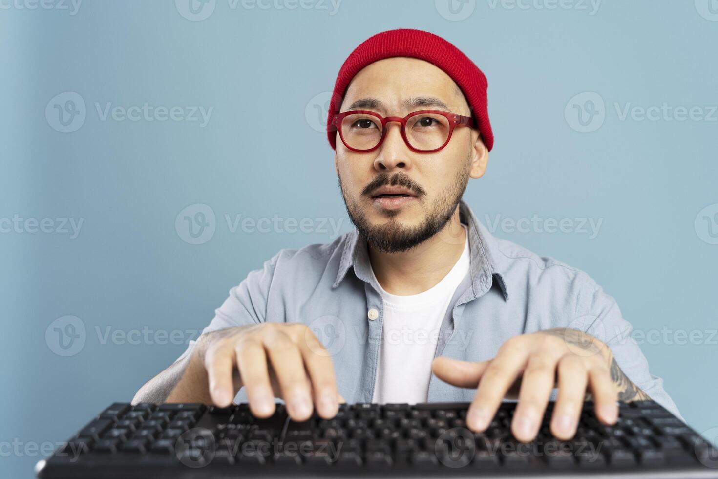 Asian man with red glasses and beanie typing on keyboard with blue background photo