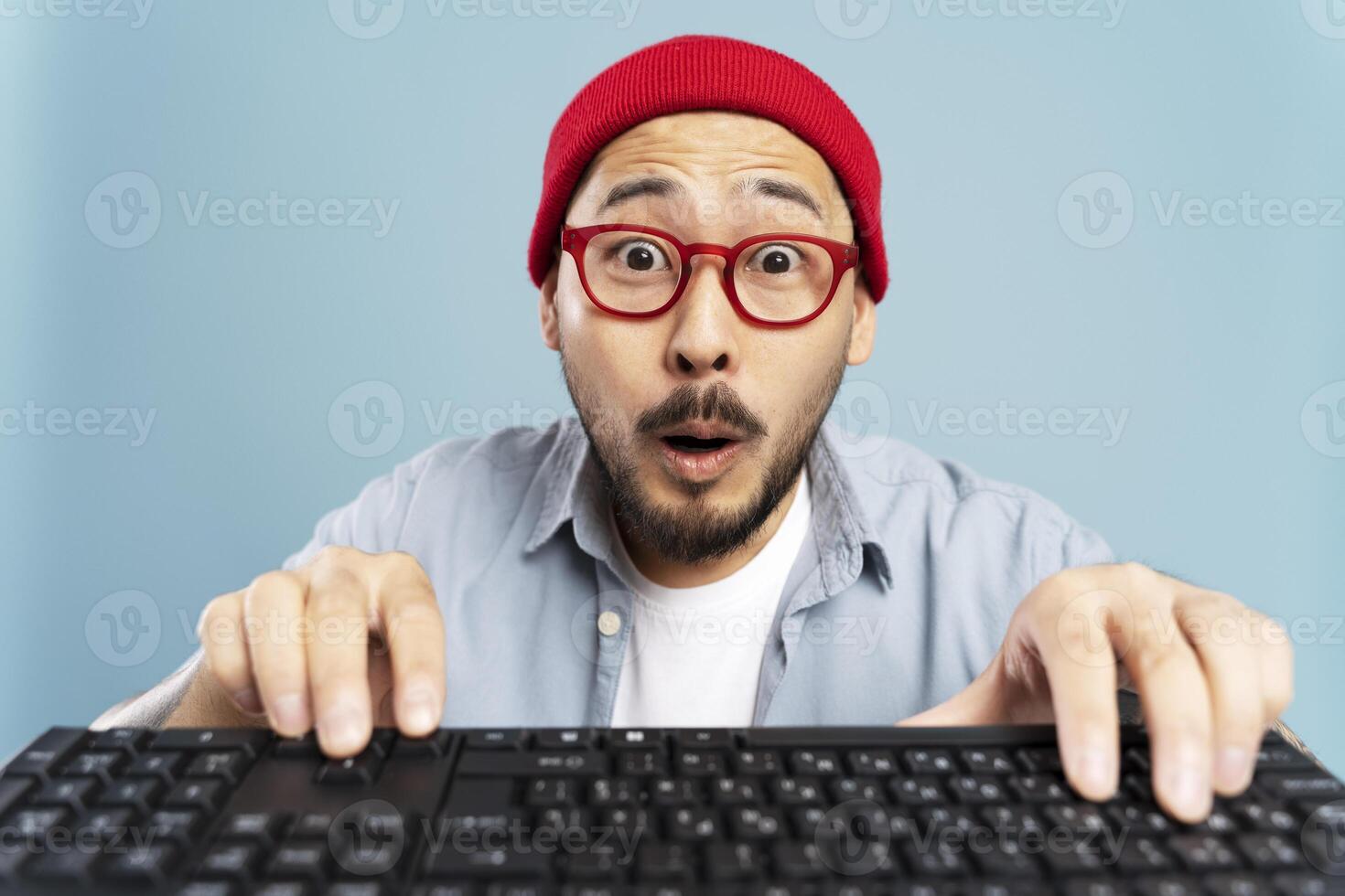 Shocked asian man wearing red glasses and beanie typing on keyboard photo