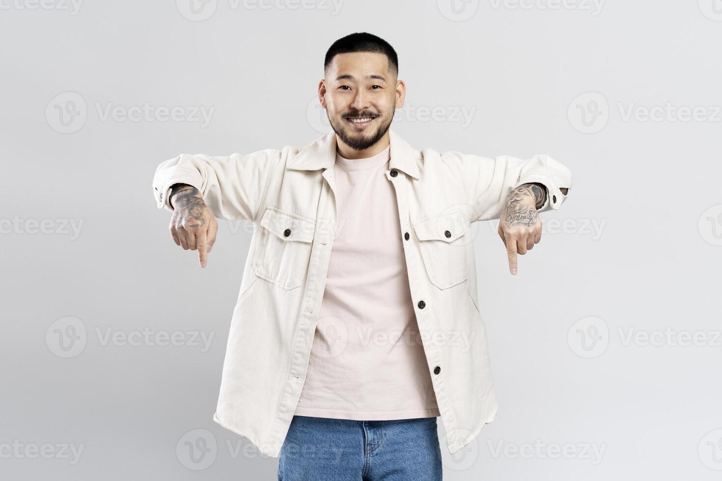 Smiling asian man with tattoos pointing down with both hands on a white background photo