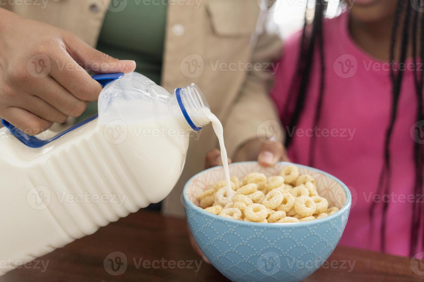 Mother pouring milk into bowl of cereal for daughter photo