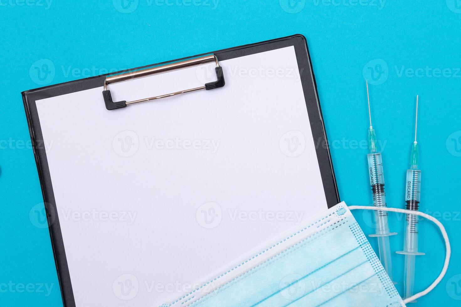 Vaccination, Immunology or Revaccination Concept - Two Medical Syringe Lying on Blue Table in Doctor's Office in a Hospital or Clinic. Black Clipboard with Sheet of Paper - Mock Up with Copy Space photo