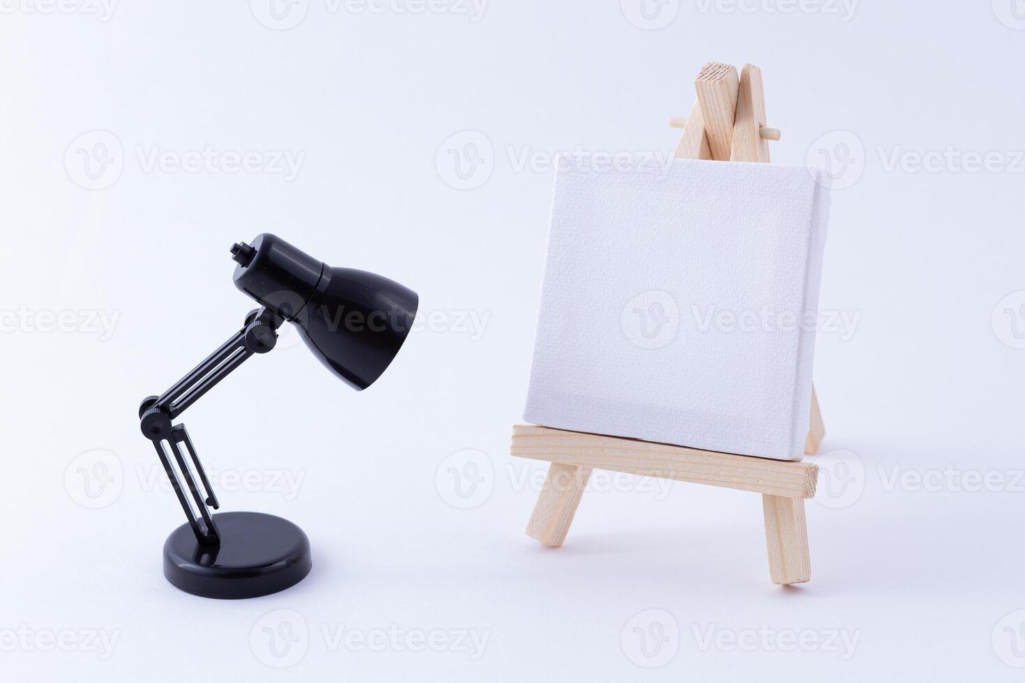 Wooden Easel Miniature with Blank White Square Canvas for Artists and Painters - Mockup. Mini Wooden Stand with Clean Artboard and Small Black Table Lamp on White Background, Copy Space photo