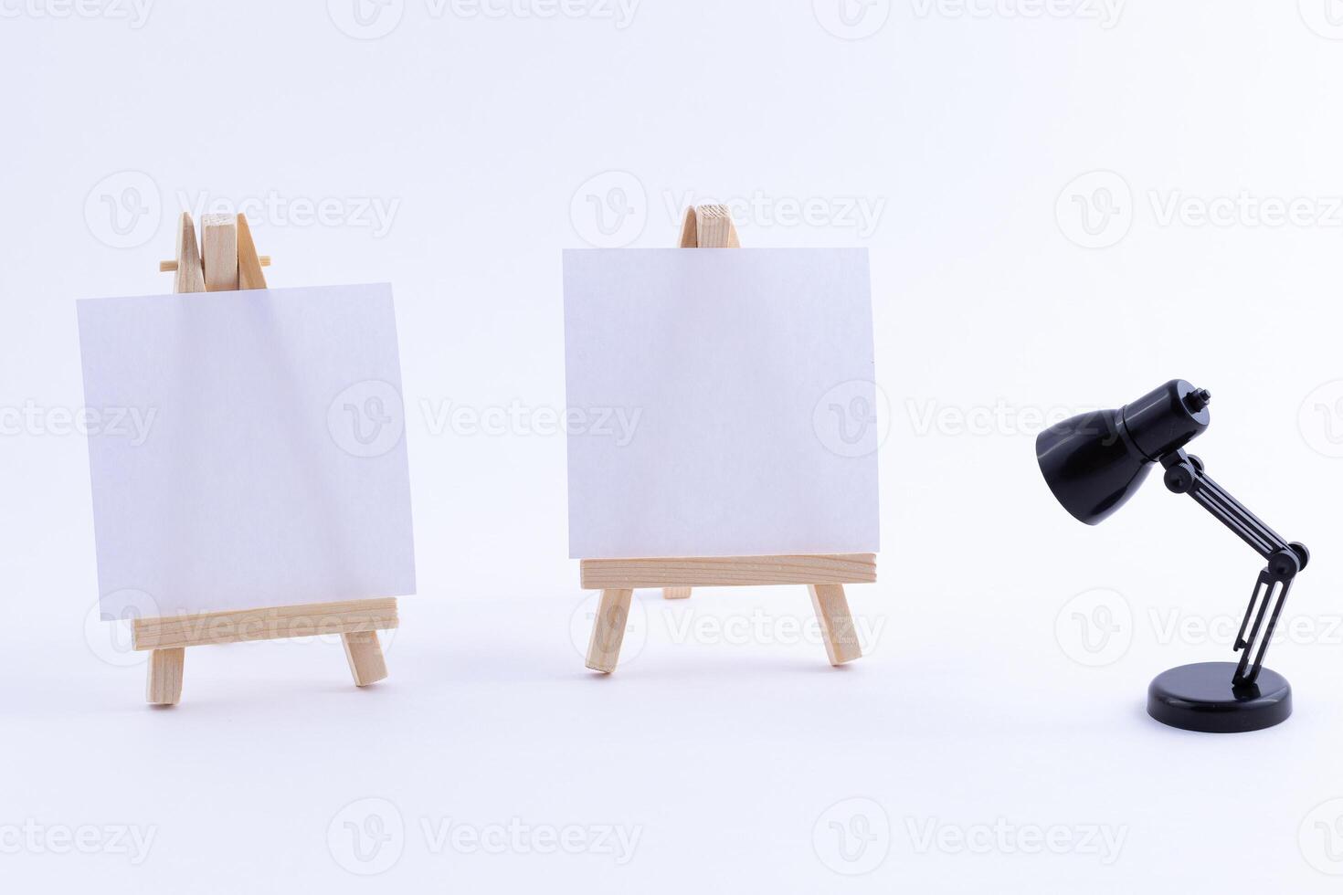 Two Wooden Easels Miniature with Blank White Square Canvas for Artists and Painters - Mockup. Mini Wooden Stands with Clean Artboard and Small Black Table Lamp on White Background, Copy Space photo