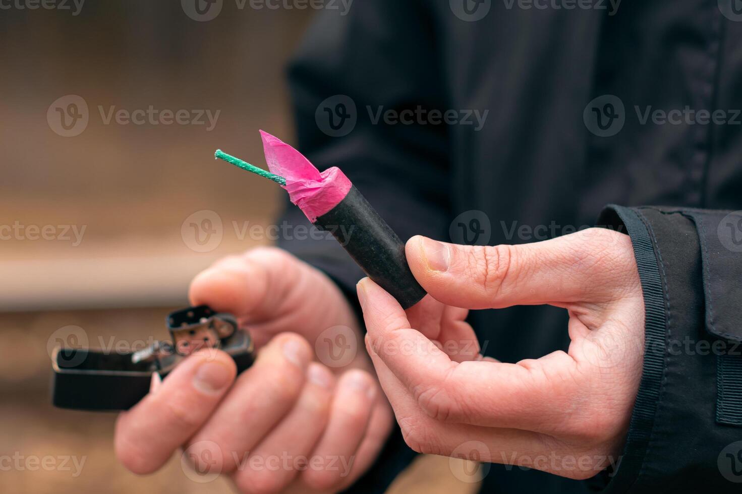 Setting Fire to the Firecracker. Man in Black Clothes Lighting Up the Petard. Firing Up the Pyrotechnics with a Black Gasoline Lighter Outdoors photo