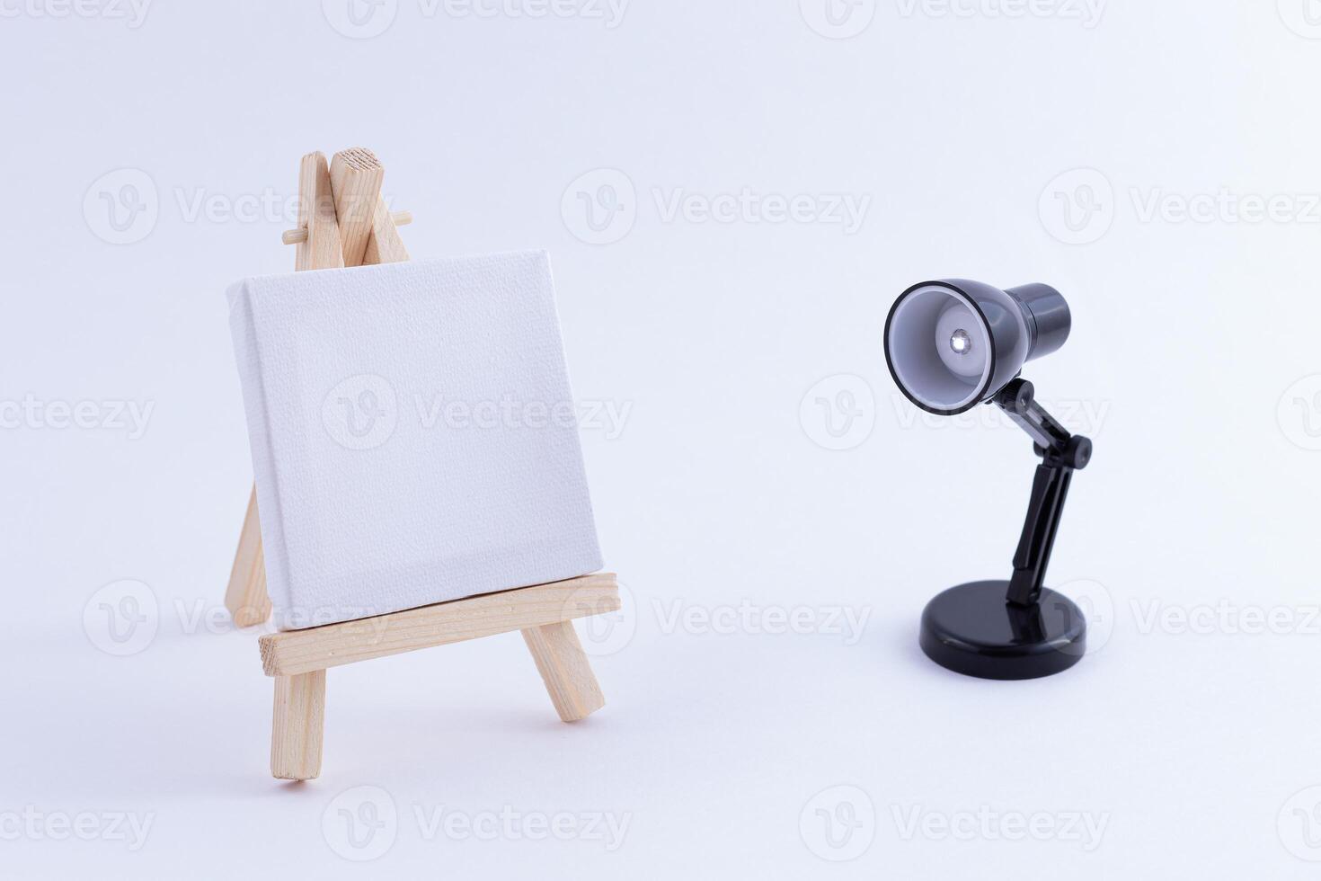 Wooden Easel Miniature with Blank White Square Canvas for Artists and Painters - Mockup. Mini Wooden Stand with Clean Artboard and Small Black Table Lamp on White Background, Copy Space photo