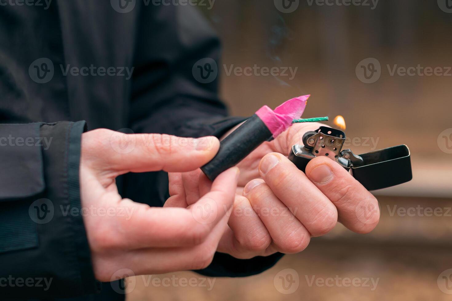 Setting Fire to the Firecracker. Man in Black Clothes Lighting Up the Petard. Firing Up the Pyrotechnics with a Black Gasoline Lighter Outdoors photo