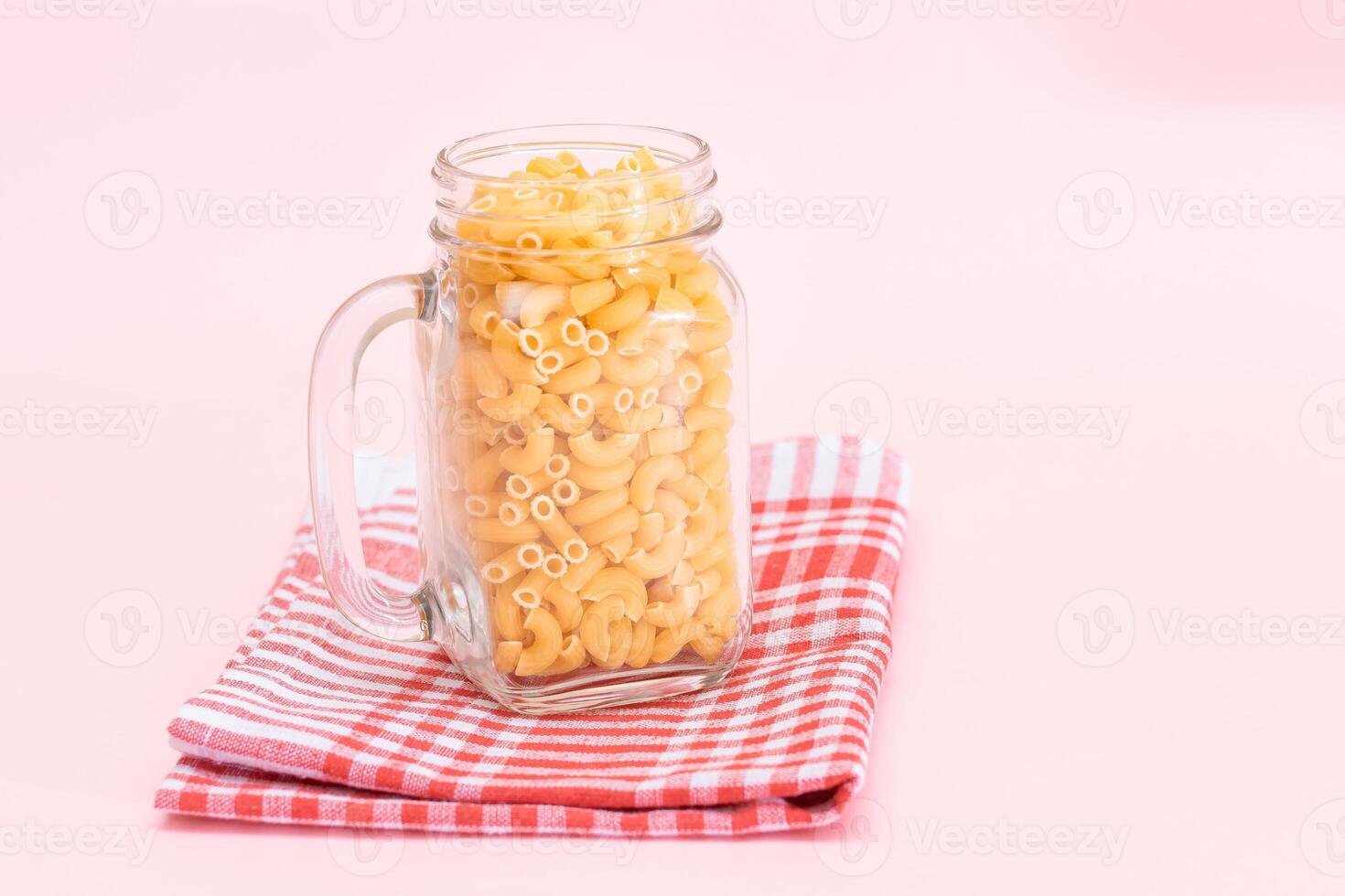 Uncooked Chifferi Rigati Pasta in Glass Jar on Checkered Red Towel on Pink Background. Fat and Unhealthy Food. Scattered Classic Dry Macaroni. Italian Culture and Cuisine. Raw Pasta photo