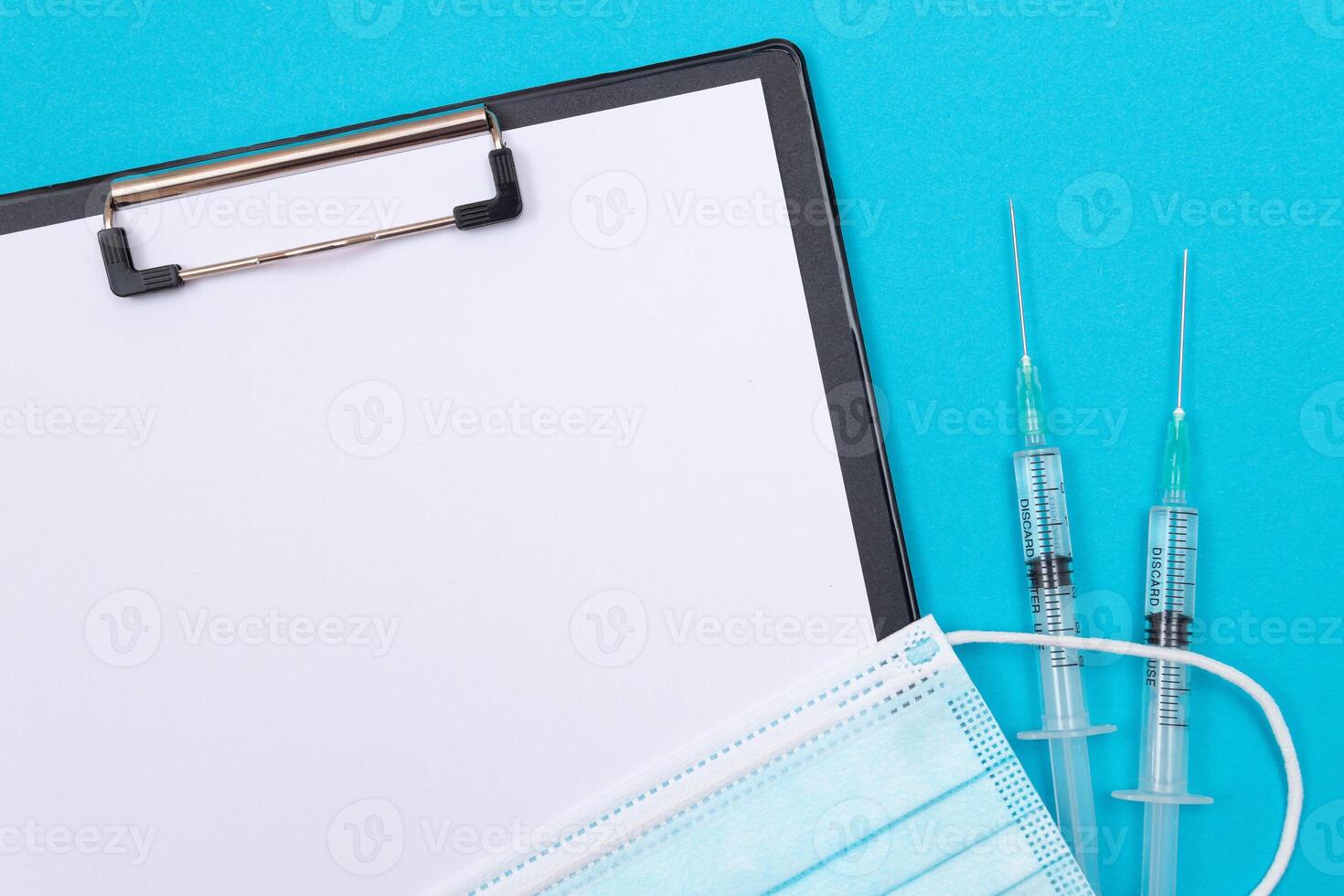 Vaccination, Immunology or Revaccination Concept - Two Medical Syringe Lying on Blue Table in Doctor's Office in a Hospital or Clinic. Black Clipboard with Sheet of Paper - Mock Up with Copy Space photo