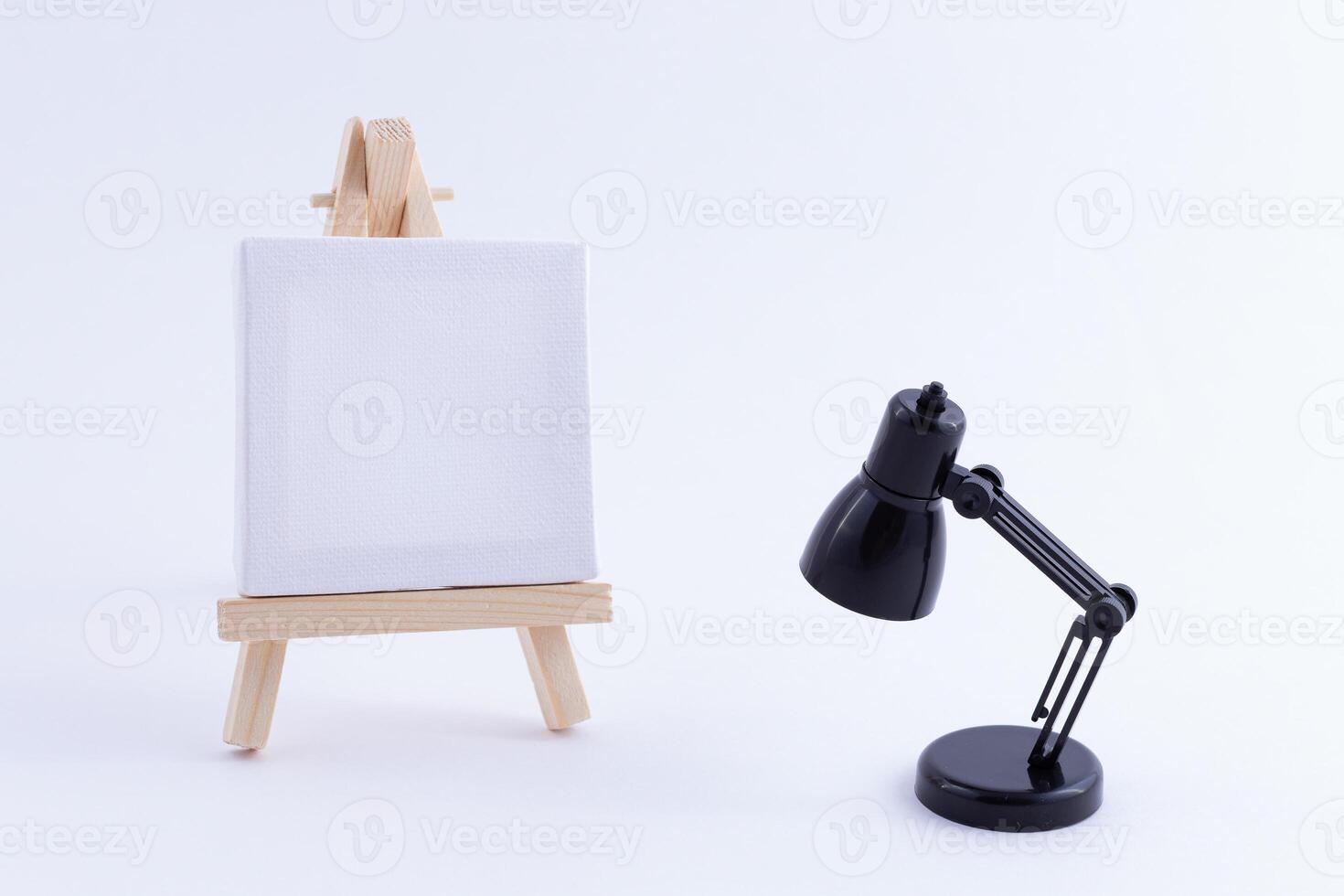 Wooden Easel Miniature with Blank White Square Canvas for Artists and Painters - Mockup. Mini Wooden Stand with Clean Artboard and Small Black Table Lamp on White Background, Copy Space photo