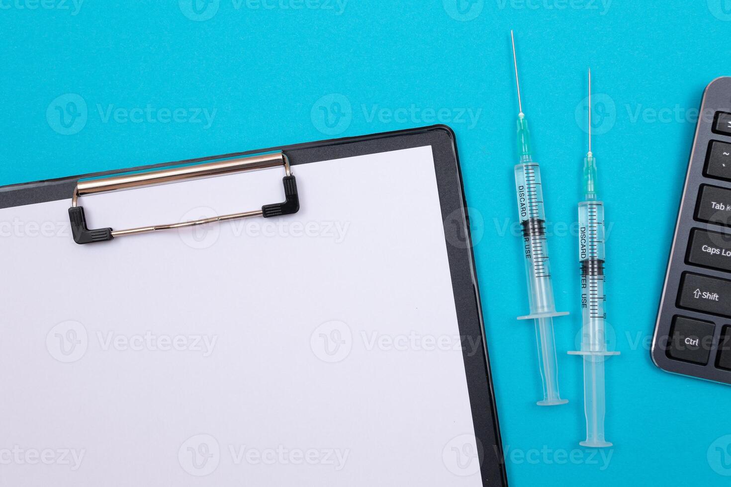 Vaccination, Immunology or Revaccination Concept - Two Medical Syringe Lying on Blue Table in Doctor's Office in a Hospital or Clinic. Black Clipboard with Sheet of Paper - Mock Up with Copy Space photo