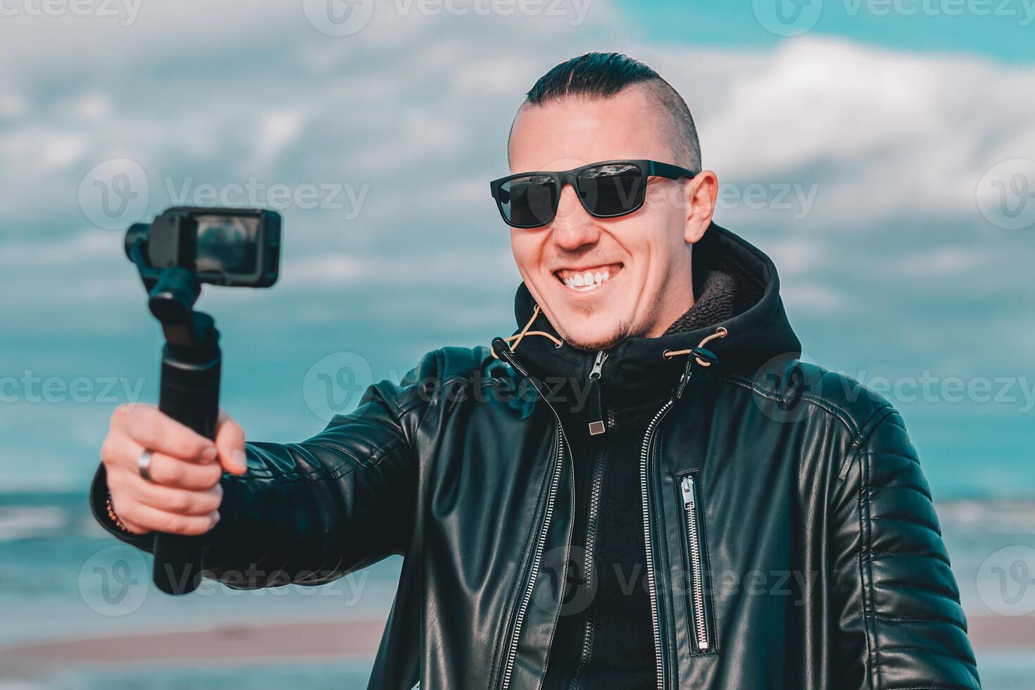 Smiling Young Male Blogger in Sunglasses Making Selfie or Streaming at the Beach Using Action Camera with Gimbal Camera Stabilizer. Hipster in Black Clothes Making Photo Against the Sea