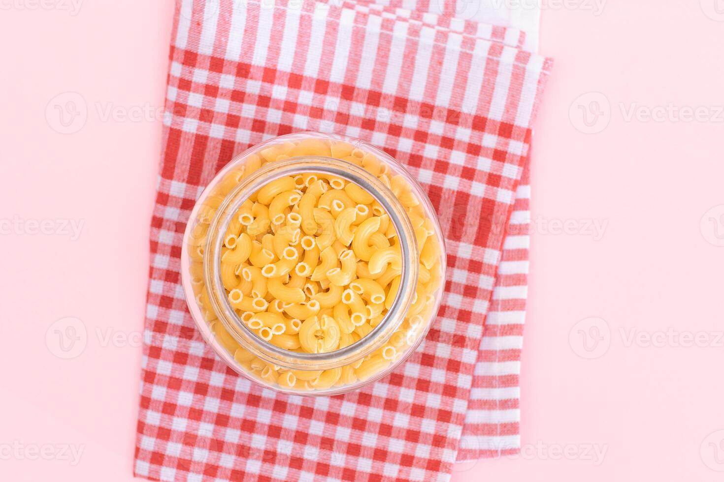 Uncooked Chifferi Rigati Pasta in Glass Jar on Checkered Red Towel on Pink Background. Fat and Unhealthy Food. Scattered Classic Dry Macaroni. Italian Culture and Cuisine. Raw Pasta photo